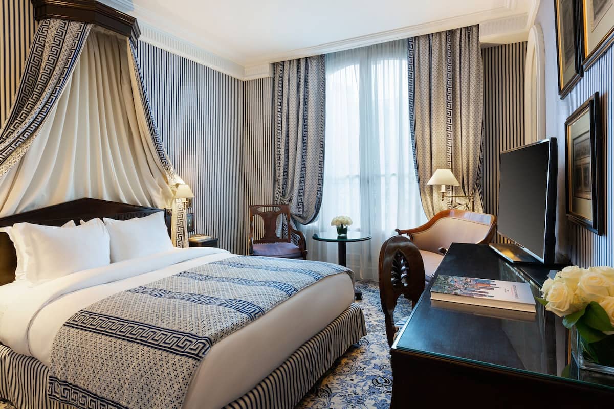 Ein Hotelzimmer mit einem großen Bett, einem Schreibtisch mit Fernseher, einem Sessel und einem kleinen Tisch mit Lampe.