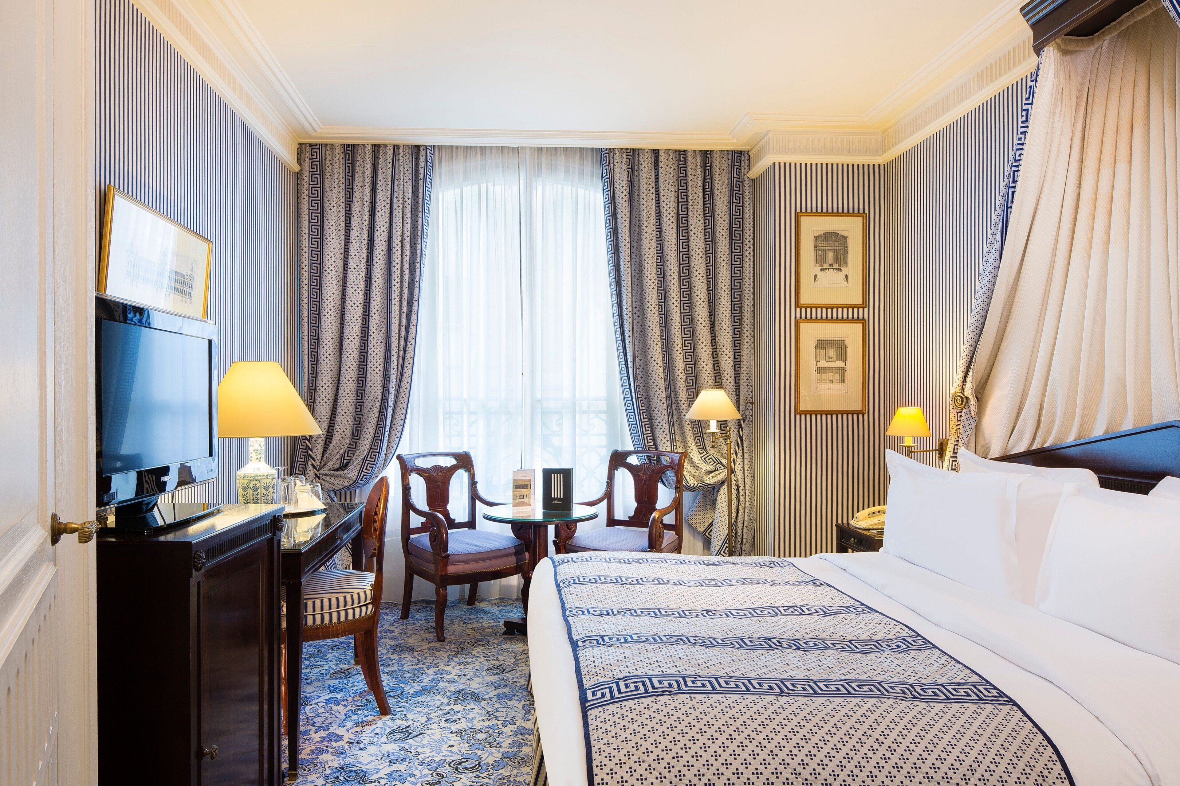 Une chambre d’hôtel avec un grand lit, un bureau avec une télévision, deux chaises, une petite table et une fenêtre avec des rideaux.