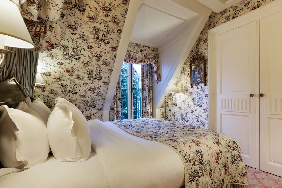Ein Schlafzimmer mit einem blumengedeckten Bett und einem Fenster mit Vorhängen. Eine weiße Tür ist ebenfalls zu sehen.