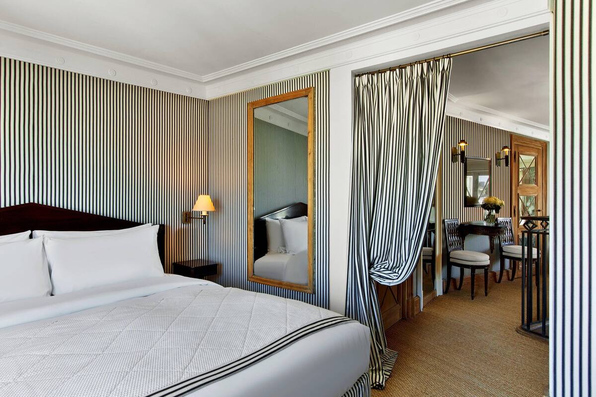 Ein Hotelzimmer mit einem großen Bett, einem Spiegel, einem Schreibtisch mit Stühlen und gestreiften Vorhängen.
