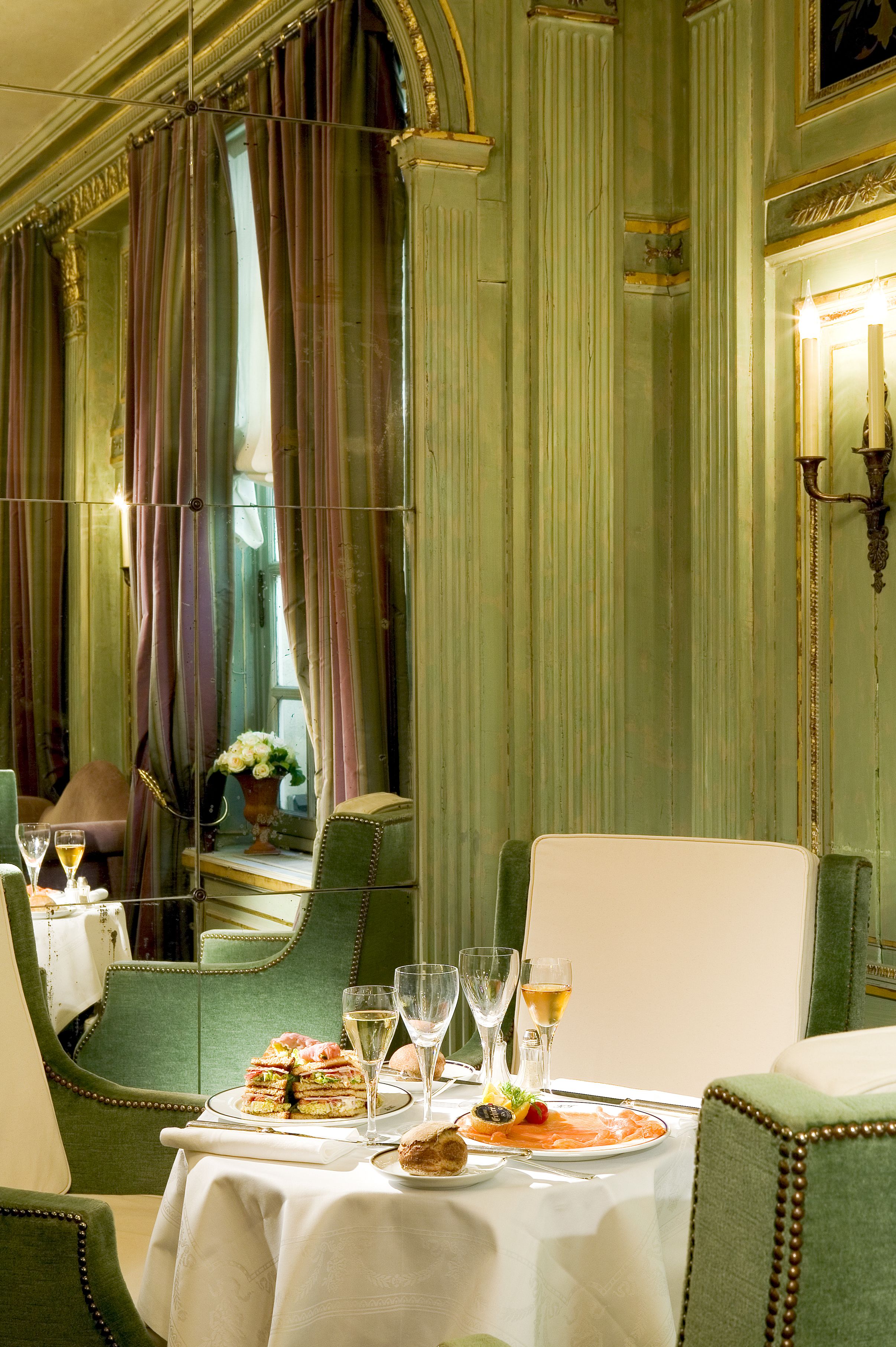 A dining table set with plates, glasses, and a vase of flowers in a room with green upholstered chairs and a mirror reflecting the room.
