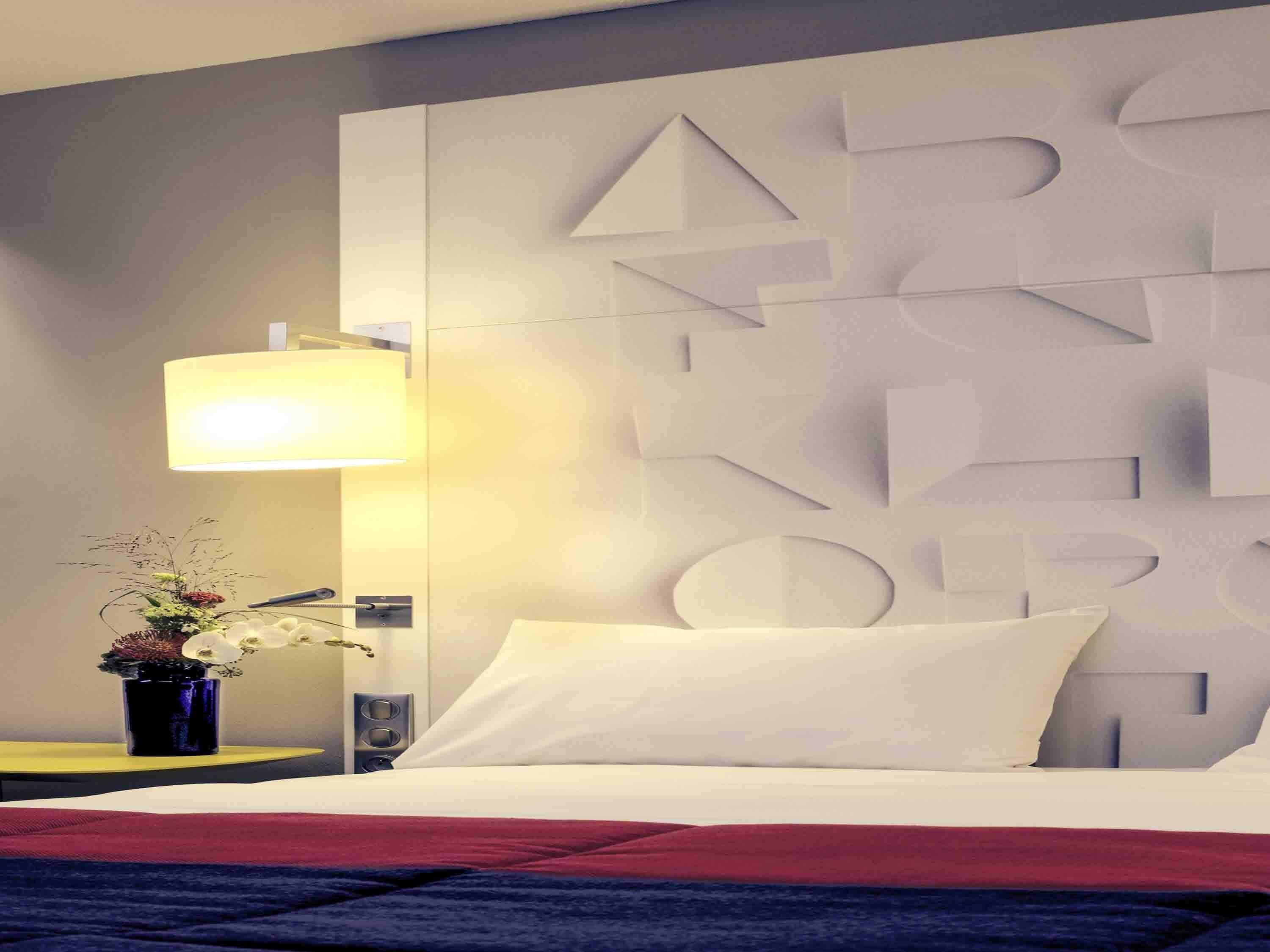 A hotel room with a bed, a wall-mounted geometric design, a bedside lamp, a small plant, and a speaker.