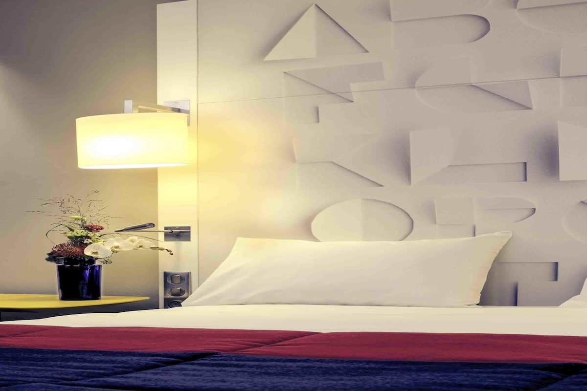 A hotel room with a bed, a wall-mounted geometric design, a bedside lamp, a small plant, and a speaker.