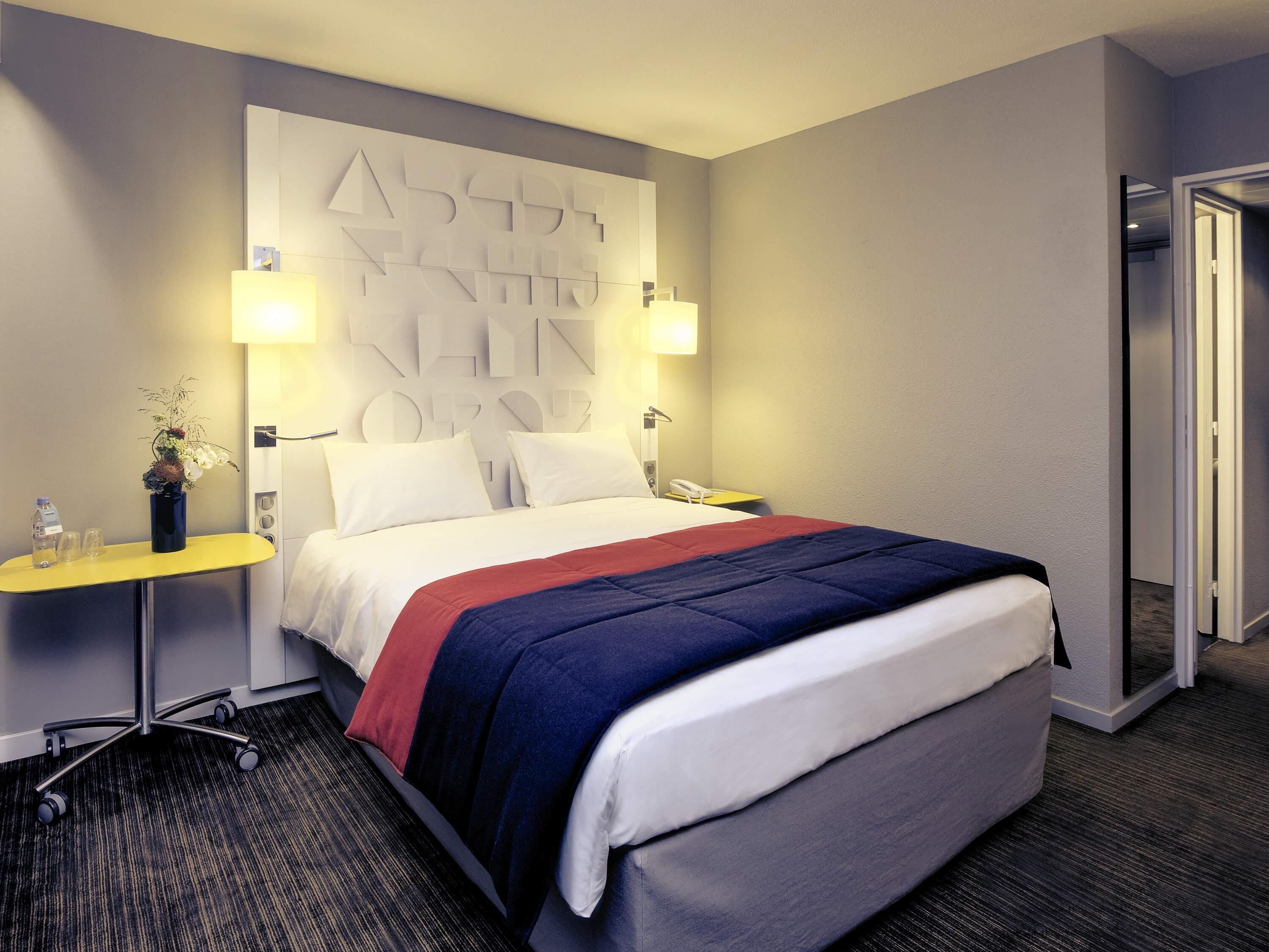 Quarto de hotel com cama, mesas de cabeceira, abajur, espelho e porta.
