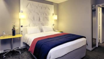 Quarto de hotel com cama, mesas de cabeceira, abajur, espelho e porta.