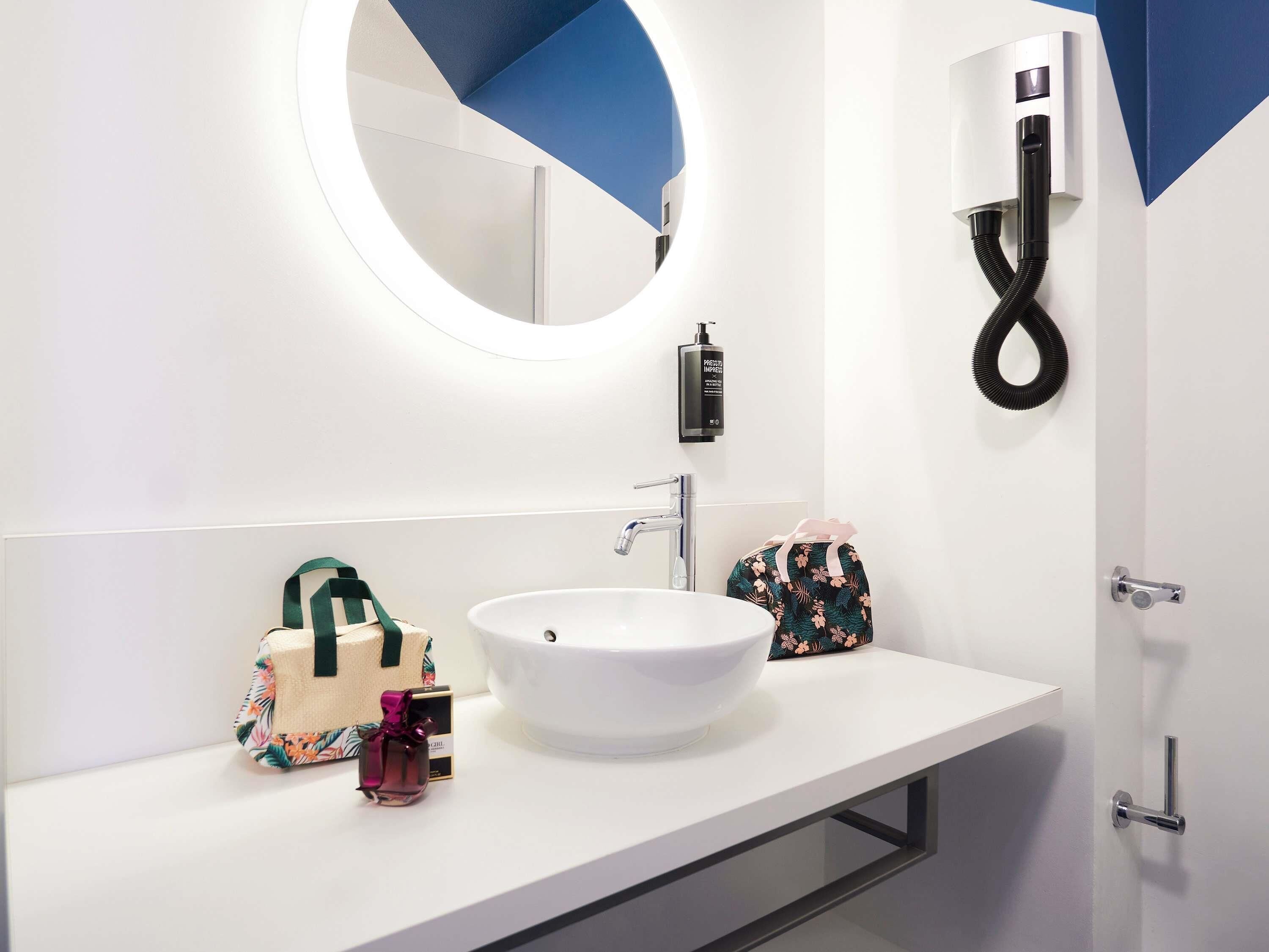 A modern bathroom with a white sink, a round mirror, a handheld showerhead, and a towel rack.