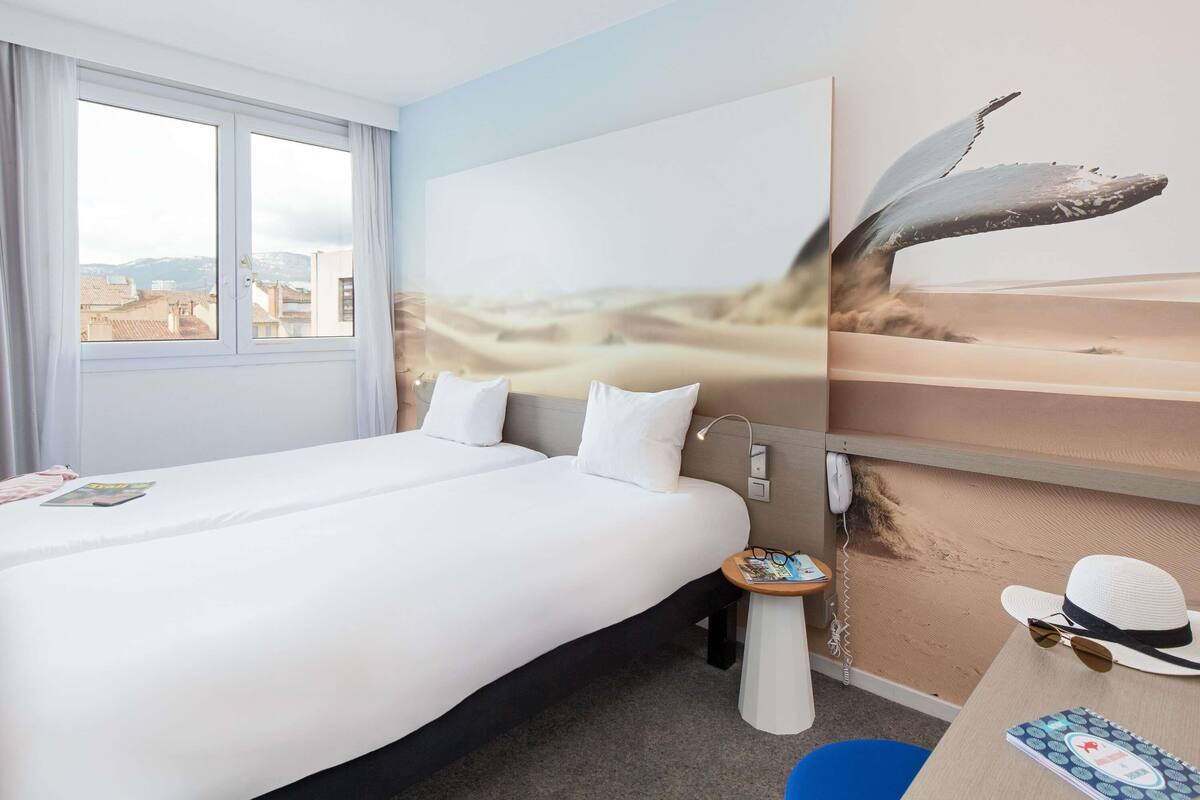 Ein Hotelzimmer mit einem großen Bett, einem kleinen Tisch, einem fest montierten Telefon und einem Fenster mit Blick auf Gebäude.