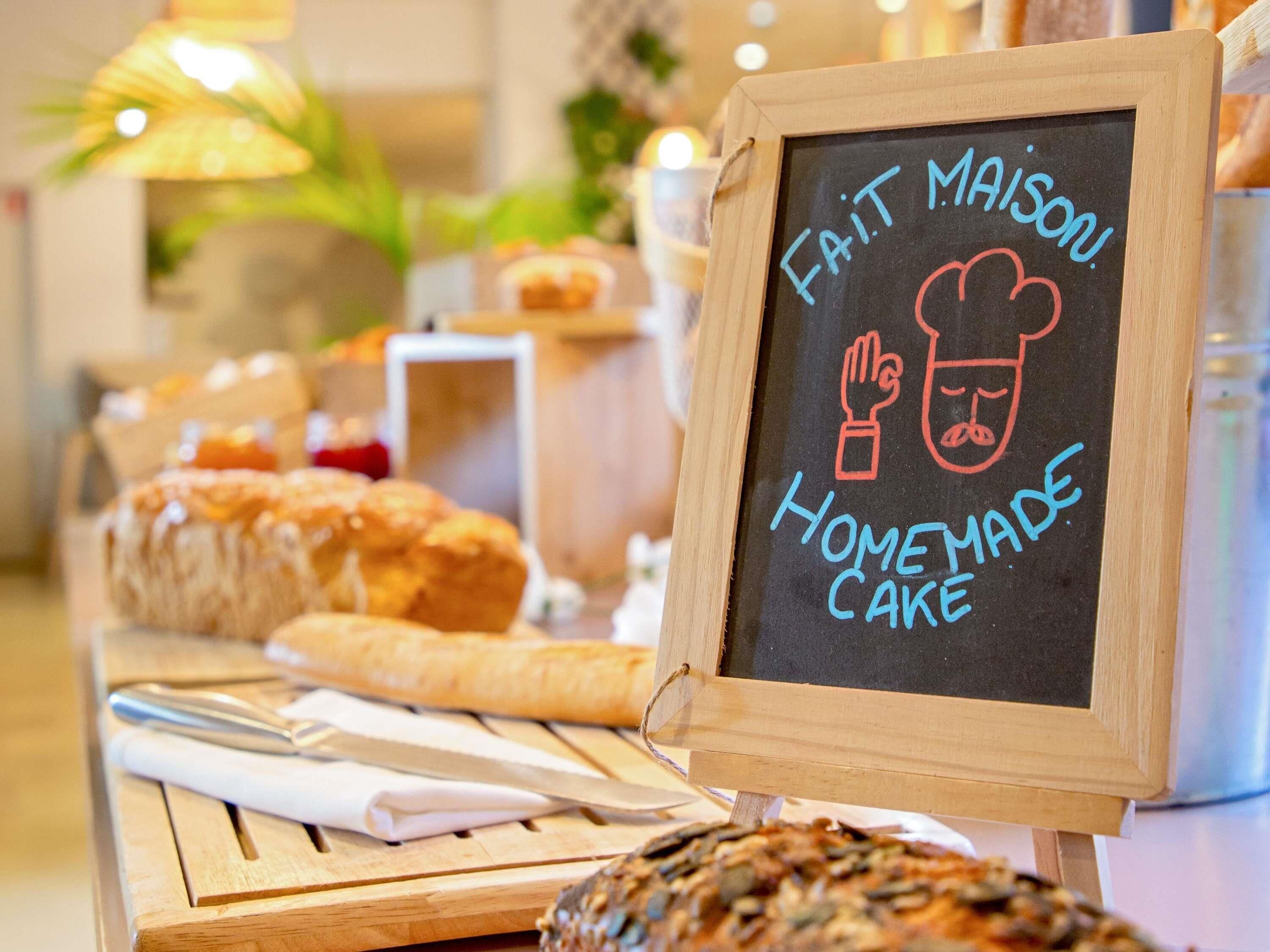 A chalkboard sign with 'FAIT MAISON Homemade Cake' written on it, placed on a wooden table with various baked goods.