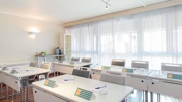 Une salle de conférence avec des tables et des chaises blanches, un projecteur et une grande fenêtre ornée de rideaux légers.
