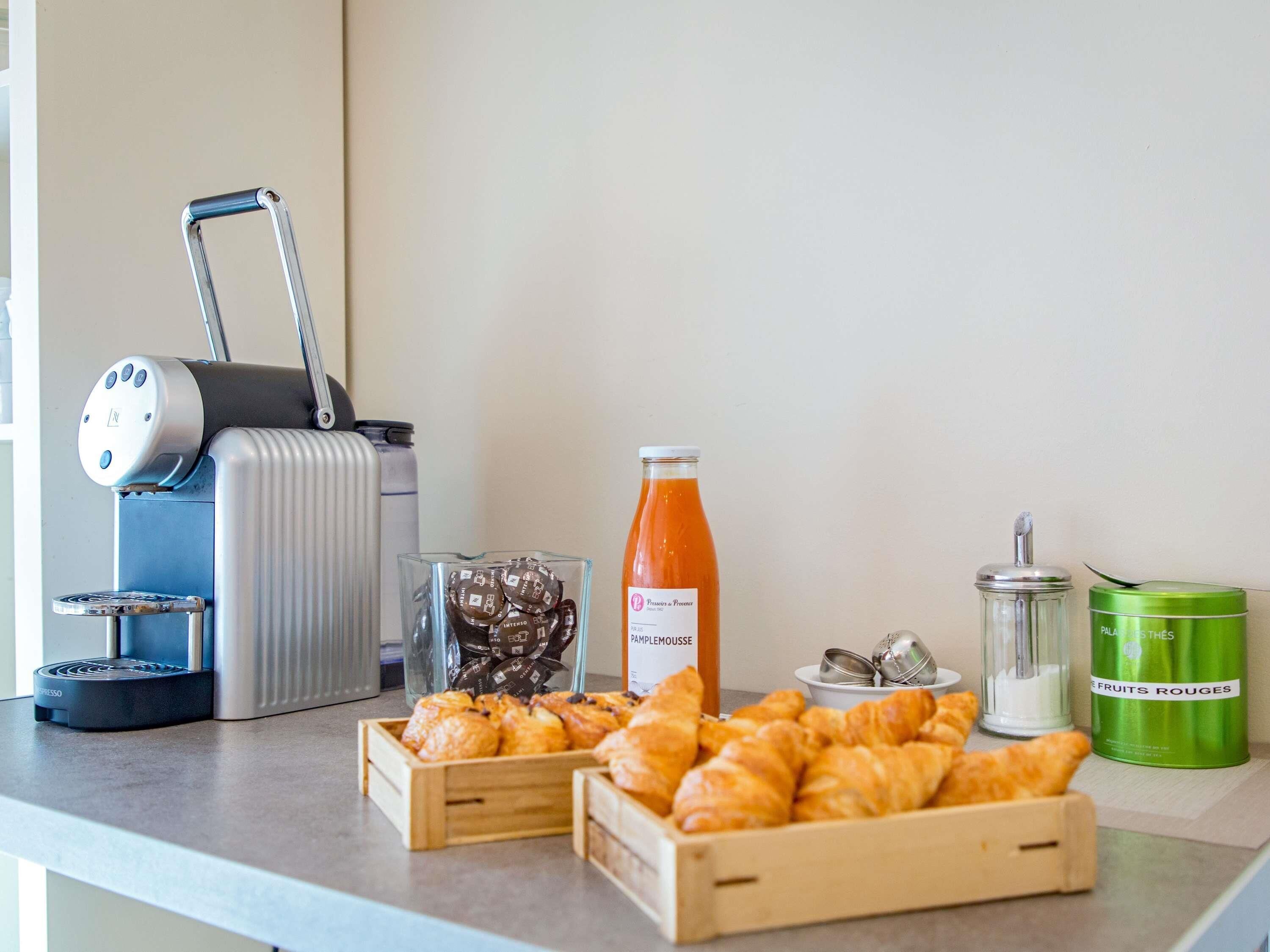 A breakfast setup with croissants, a juice bottle, a coffee machine, and a green container of red fruits.