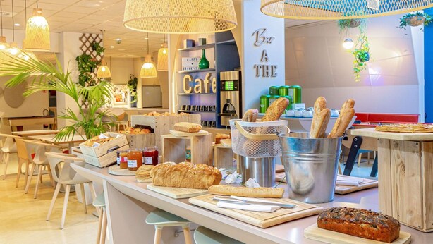 Un café avec un comptoir où sont exposés du pain et des pâtisseries, une machine à café et des lampes en osier suspendues au plafond.