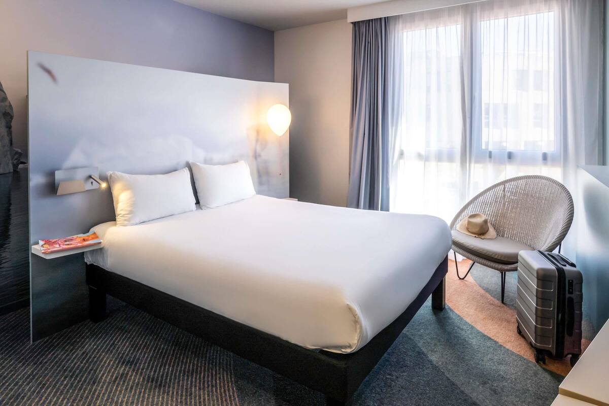 Ein Hotelzimmer mit einem großen Bett, einem Nachttisch, einem Sessel, einem Koffer und einem Fenster mit Vorhängen.