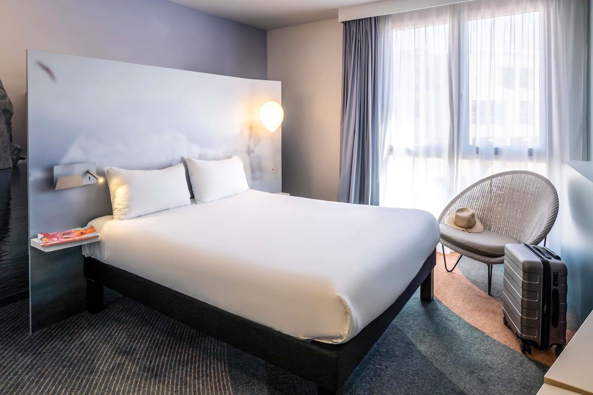 A hotel room with a large bed, a nightstand, a chair, a suitcase, and a window with curtains.