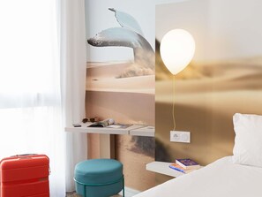 Een hotelkamer met een grote muurschildering van een walvis, een nachtkastje met boeken, een rode koffer, een blauwgroen voetenbankje en een wandlamp.