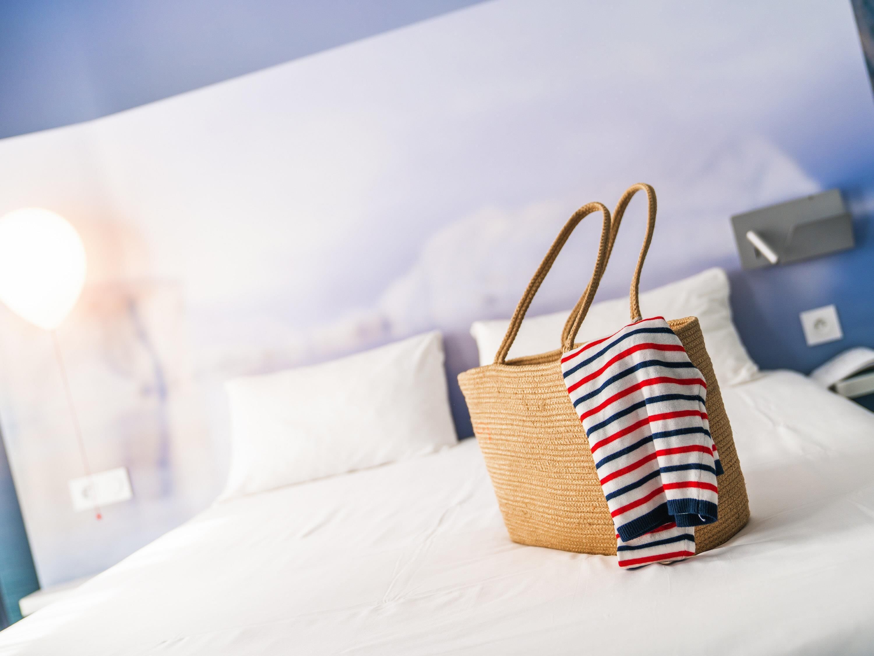 A bed with white bedding and a straw-colored woven bag with a striped towel.