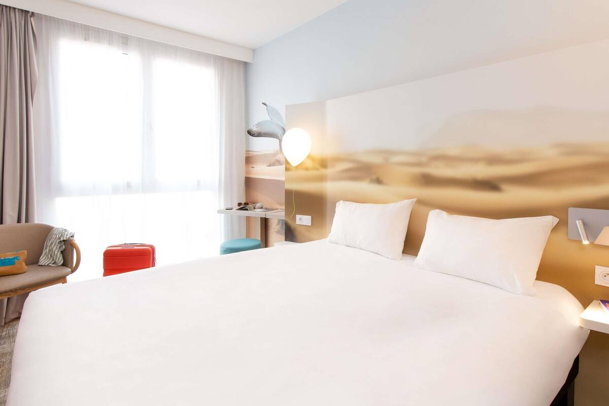 Ein Hotelzimmer mit einem großen Bett, einem Schreibtisch, einem Stuhl, einem Koffer und einem Fenster mit Vorhängen.