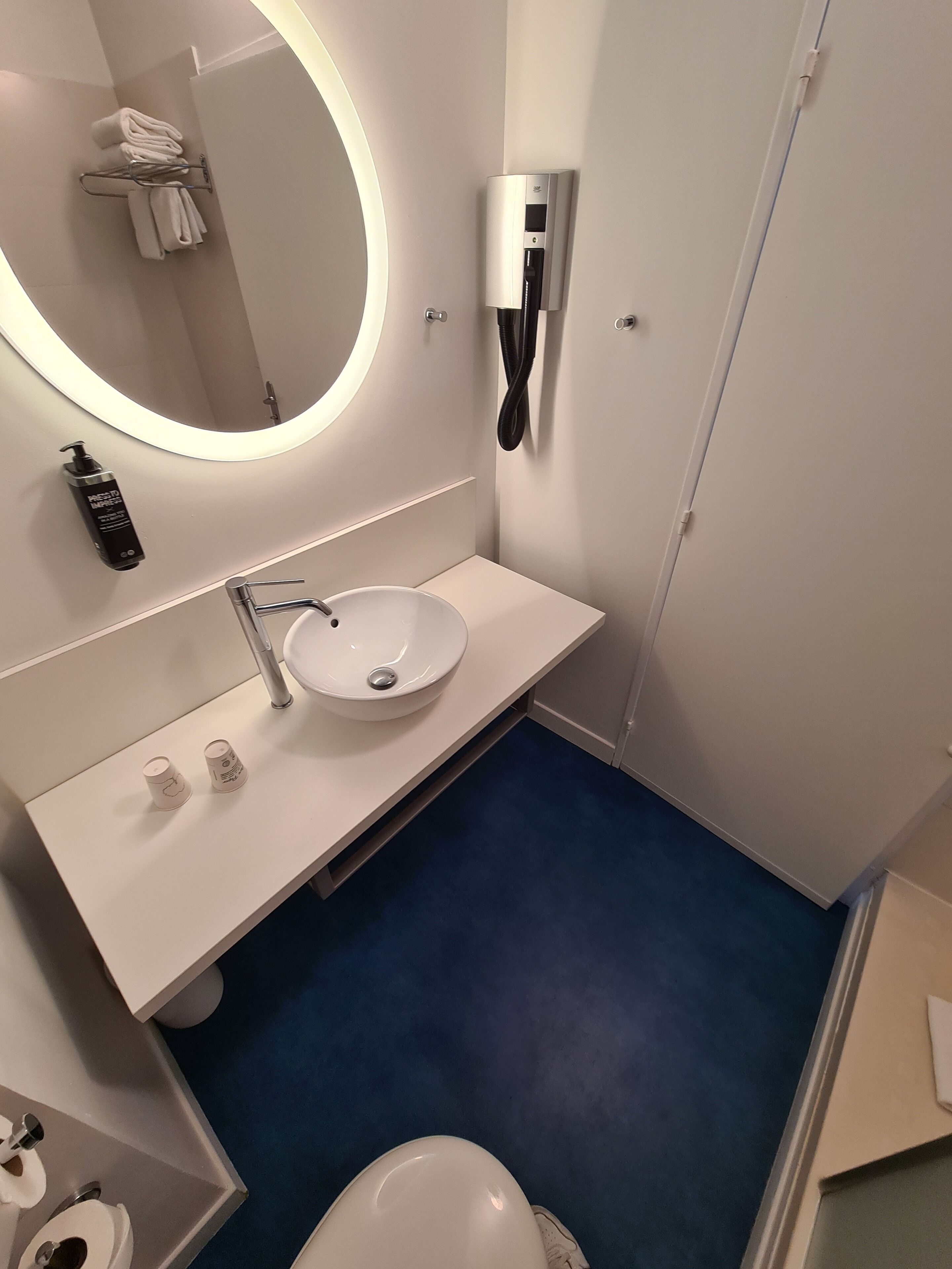 A modern bathroom with a white sink, a round mirror, a towel rack, and a hand dryer.