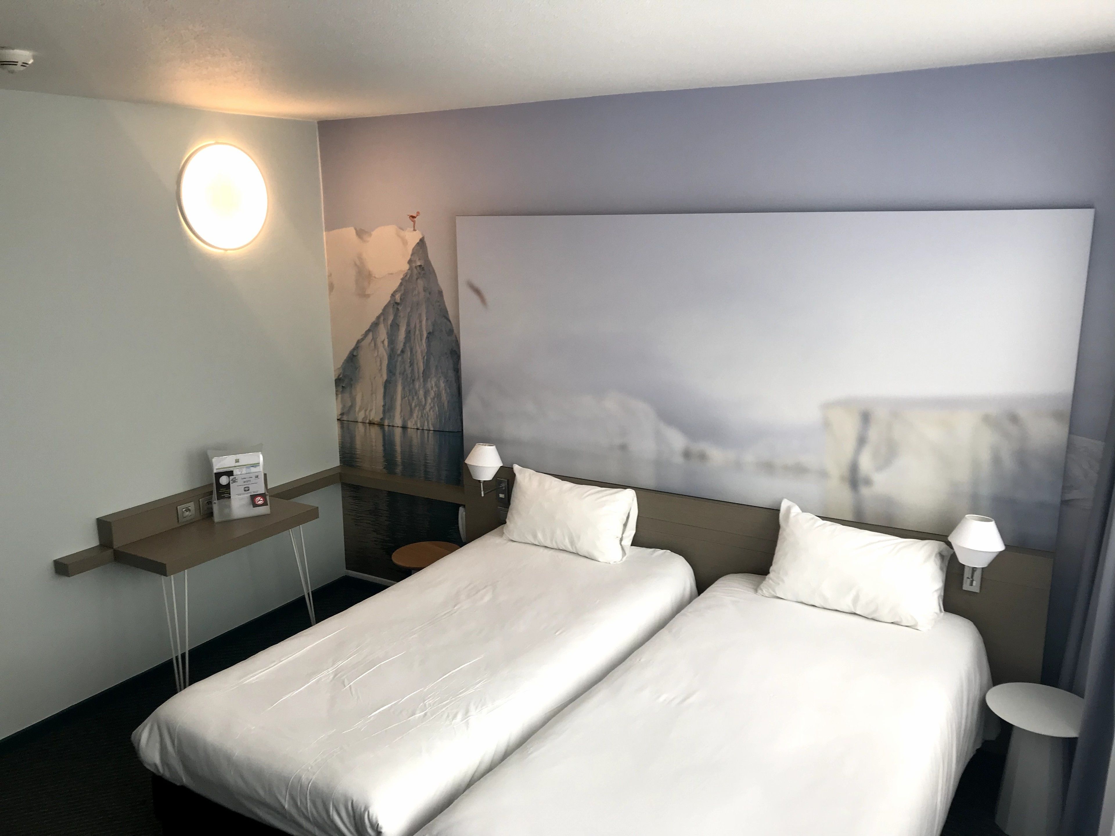 Une chambre d’hôtel avec deux lits, une lampe fixée au mur, un tableau représentant une montagne et une petite table avec une lampe.