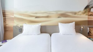 Une chambre d’hôtel avec deux lits, une grande peinture murale représentant un désert et une petite table sur laquelle se trouve un livre.