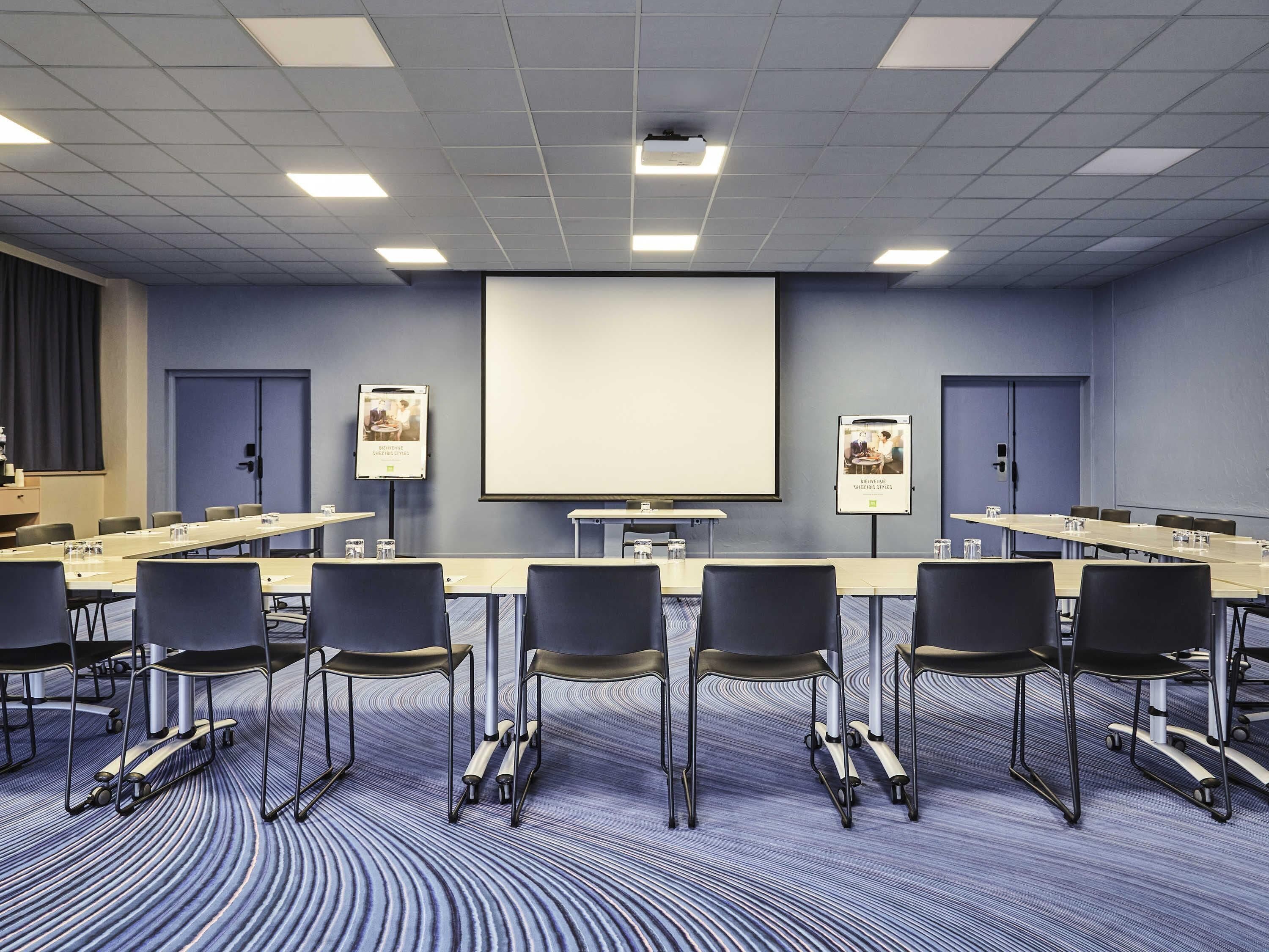 Una sala de conferències amb una pantalla gran, diverses taules, cadires i un sòl amb moqueta.