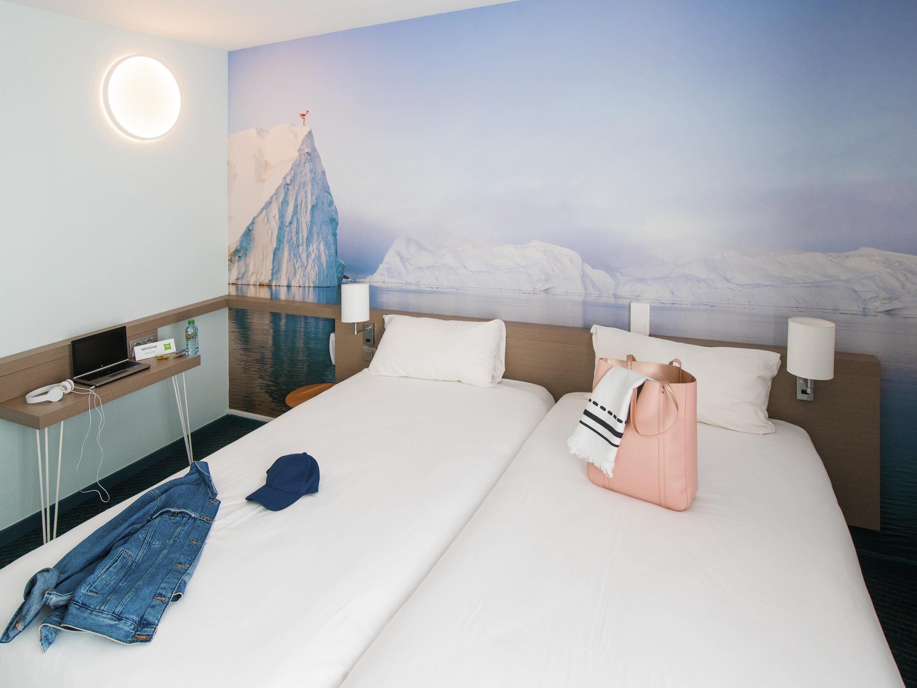 A hotel room with a bed, a desk with a laptop, a chair, a wall-mounted lamp, and a painting of a mountain.