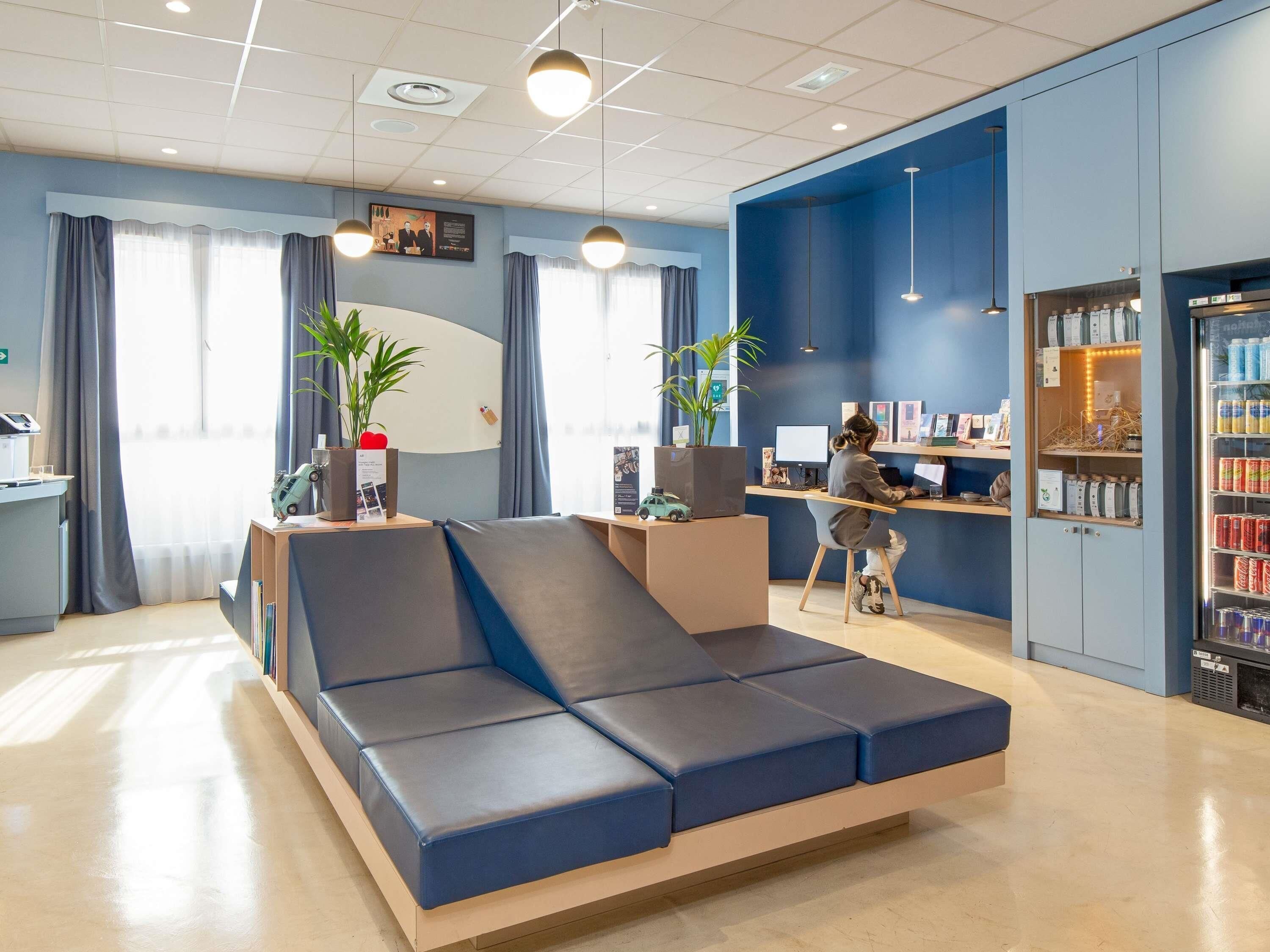 Un hall d’hôtel moderne avec un canapé bleu, un comptoir d’accueil, un poste informatique et un réfrigérateur pour les boissons.