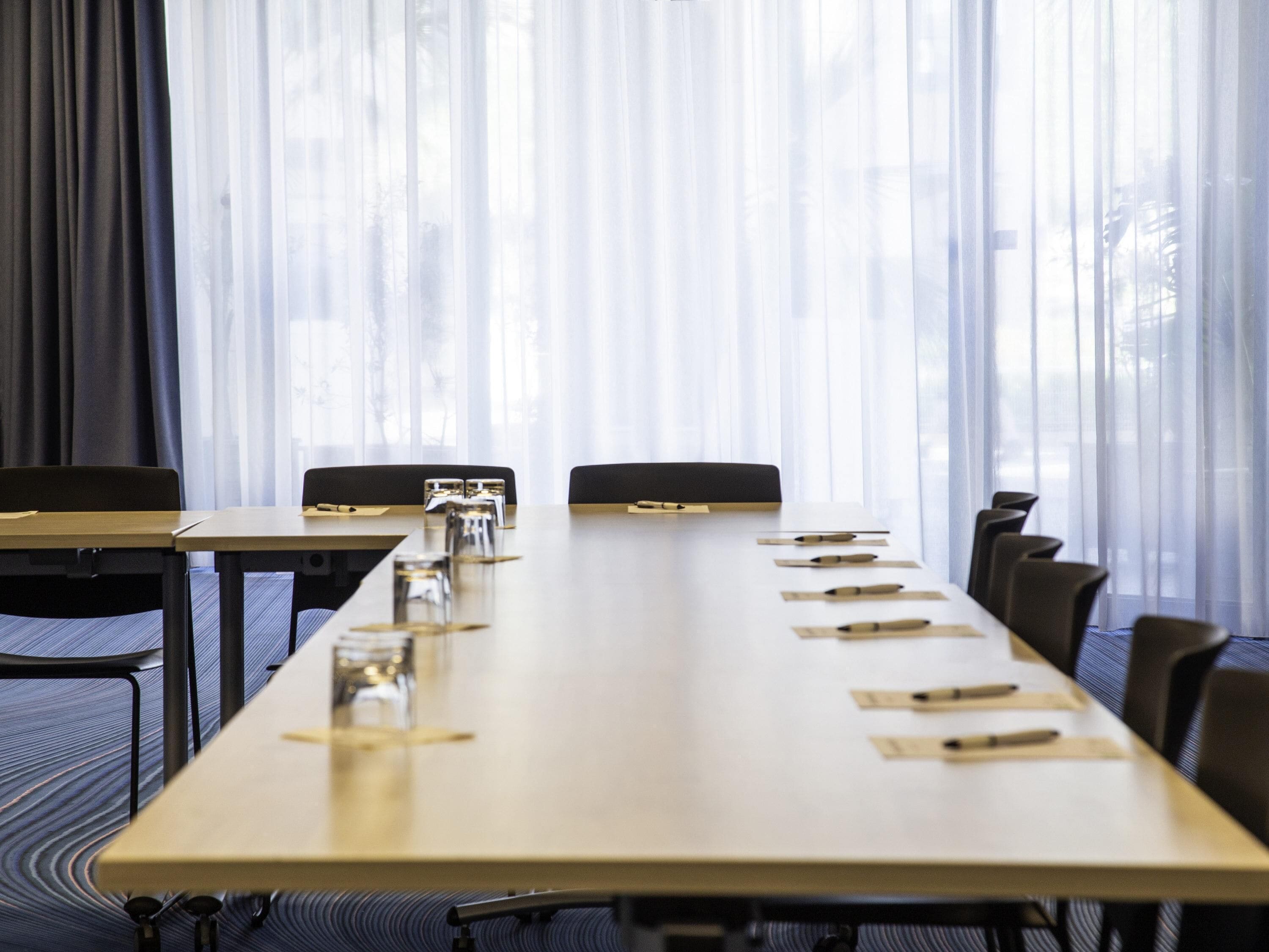 A conference room with a long table, chairs, and a window with sheer curtains.