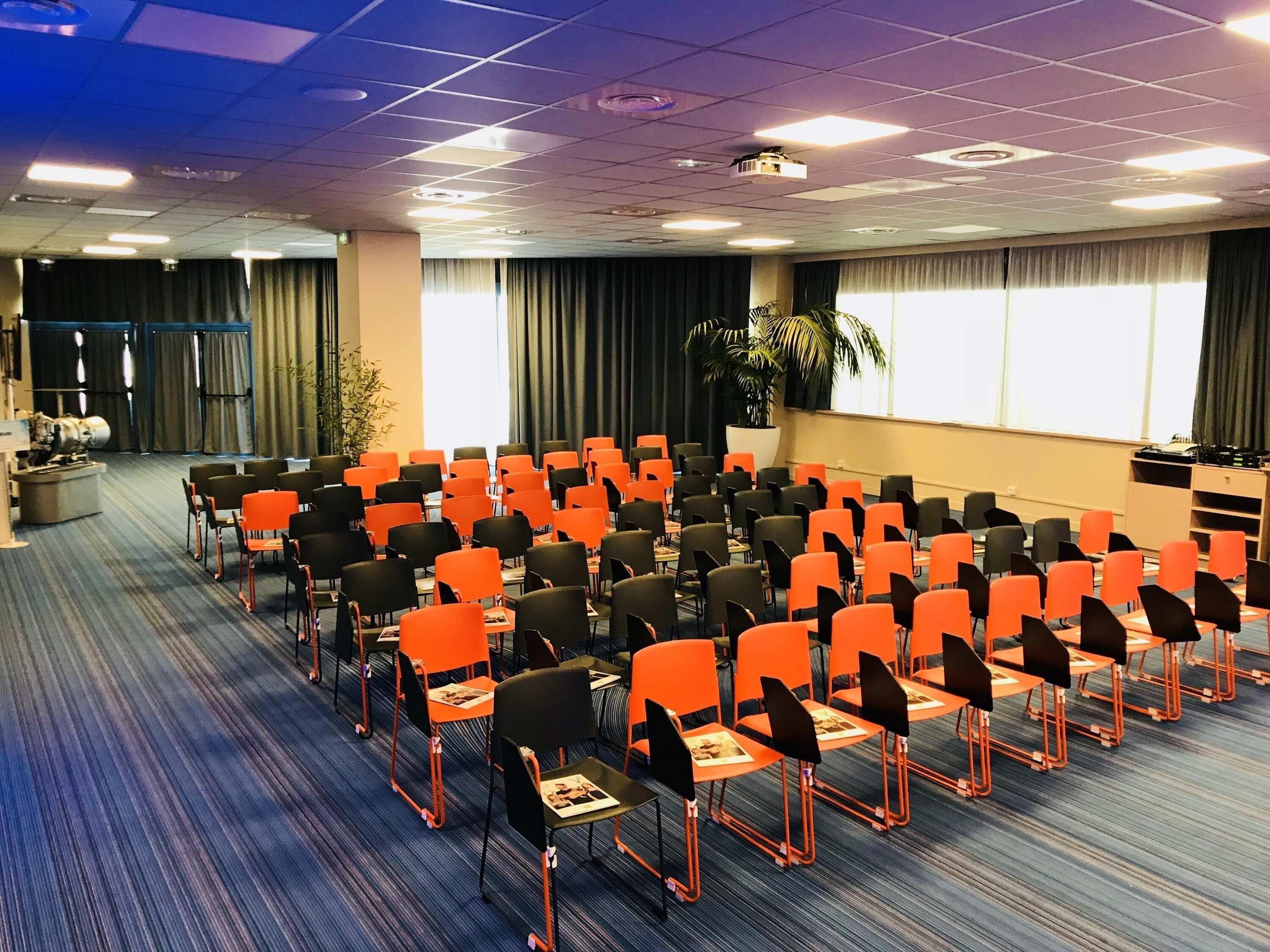 Un salón de conferencias con filas de sillas, un escenario, cortinas y un proyector.