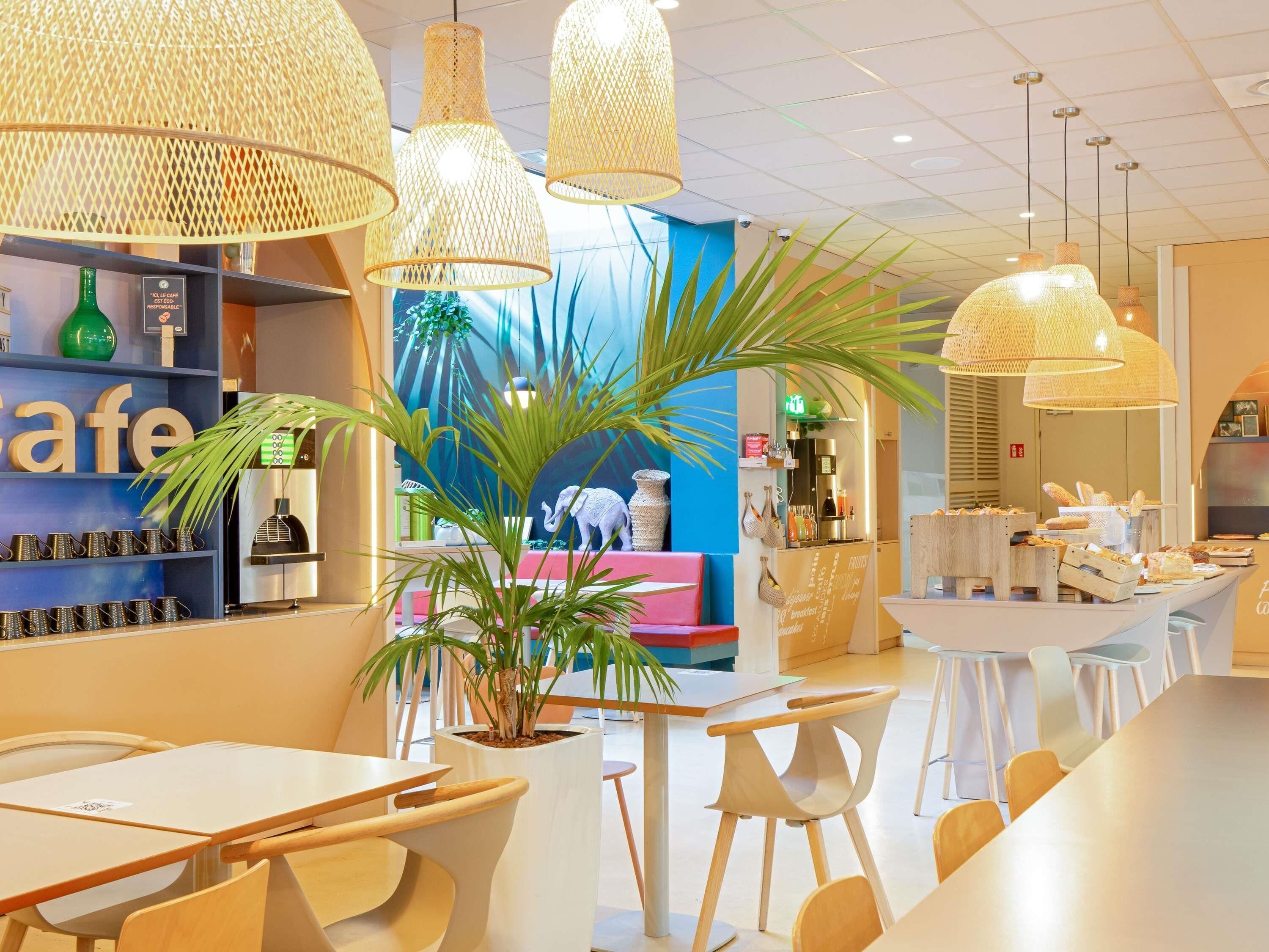 Interior de una cafetería moderna con lámparas de mimbre colgantes, un mostrador que exhibe productos alimenticios y una zona de estar con mesas y sillas.