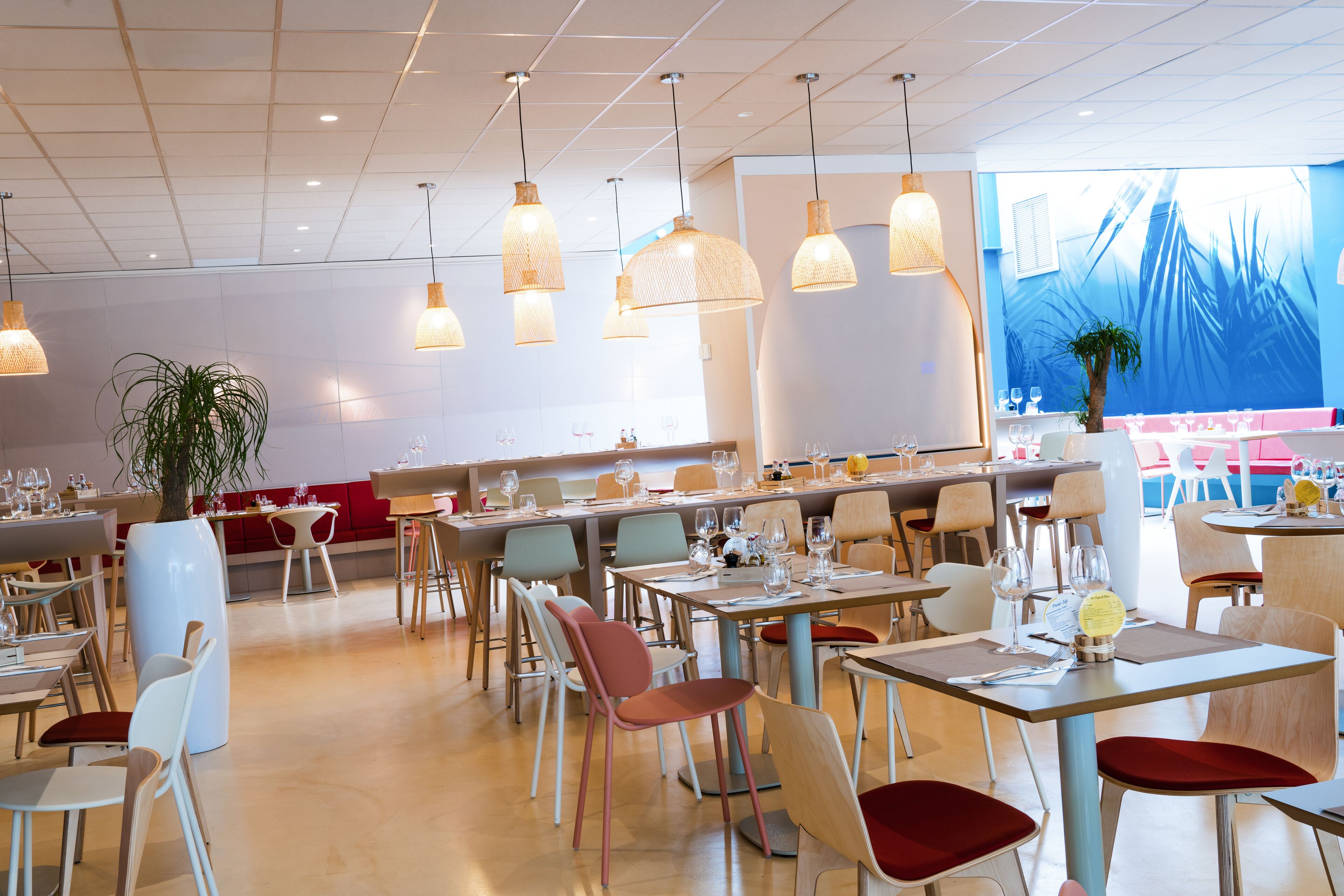 A spacious restaurant with multiple dining tables and chairs, hanging pendant lights, and a large window with a view of palm trees.