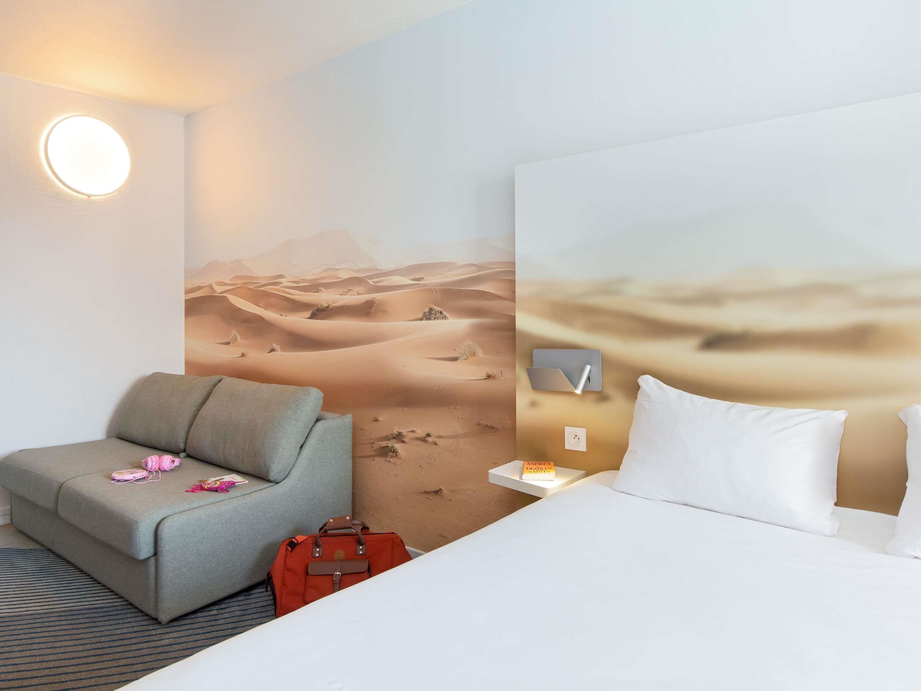 A hotel room with a bed, a grey armchair, a red suitcase, a wall-mounted lamp, and a desert-themed mural.