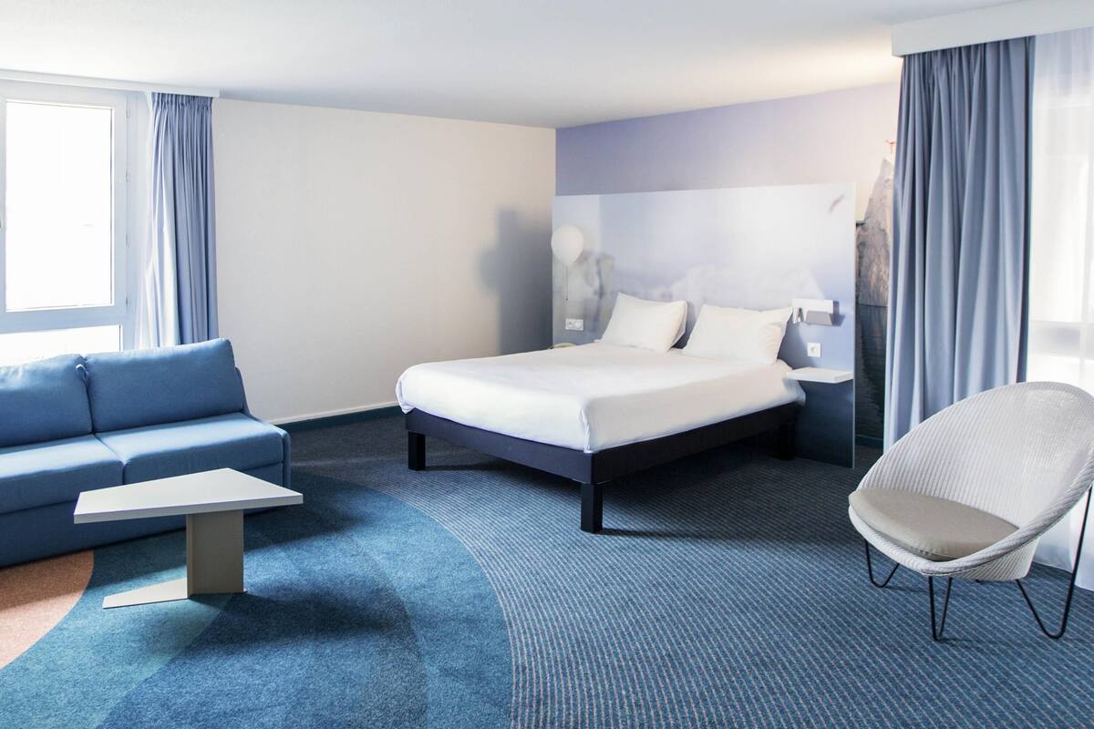 Ein Hotelzimmer mit Bett, Sofa, Sessel, kleinem Tisch und einem Fenster mit Vorhängen.