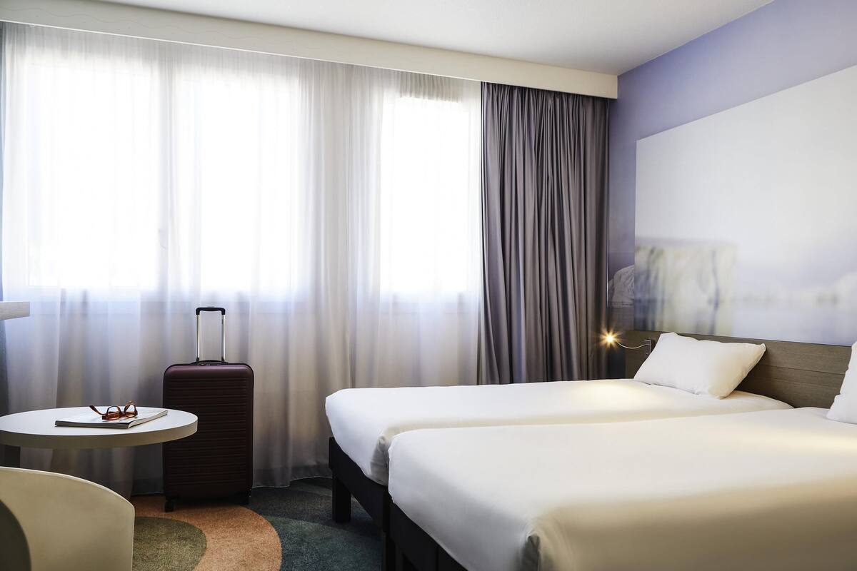 Ein Hotelzimmer mit zwei Betten, einem kleinen Tisch und einem Koffer, sowie einem großen Fenster mit Vorhängen.