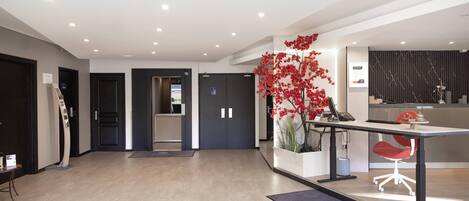 A modern hotel lobby with a reception desk, a red chair, a potted plant, and a digital screen.