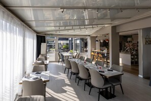A modern dining area with tables and chairs, a kitchen in the background, and large windows allowing natural light.