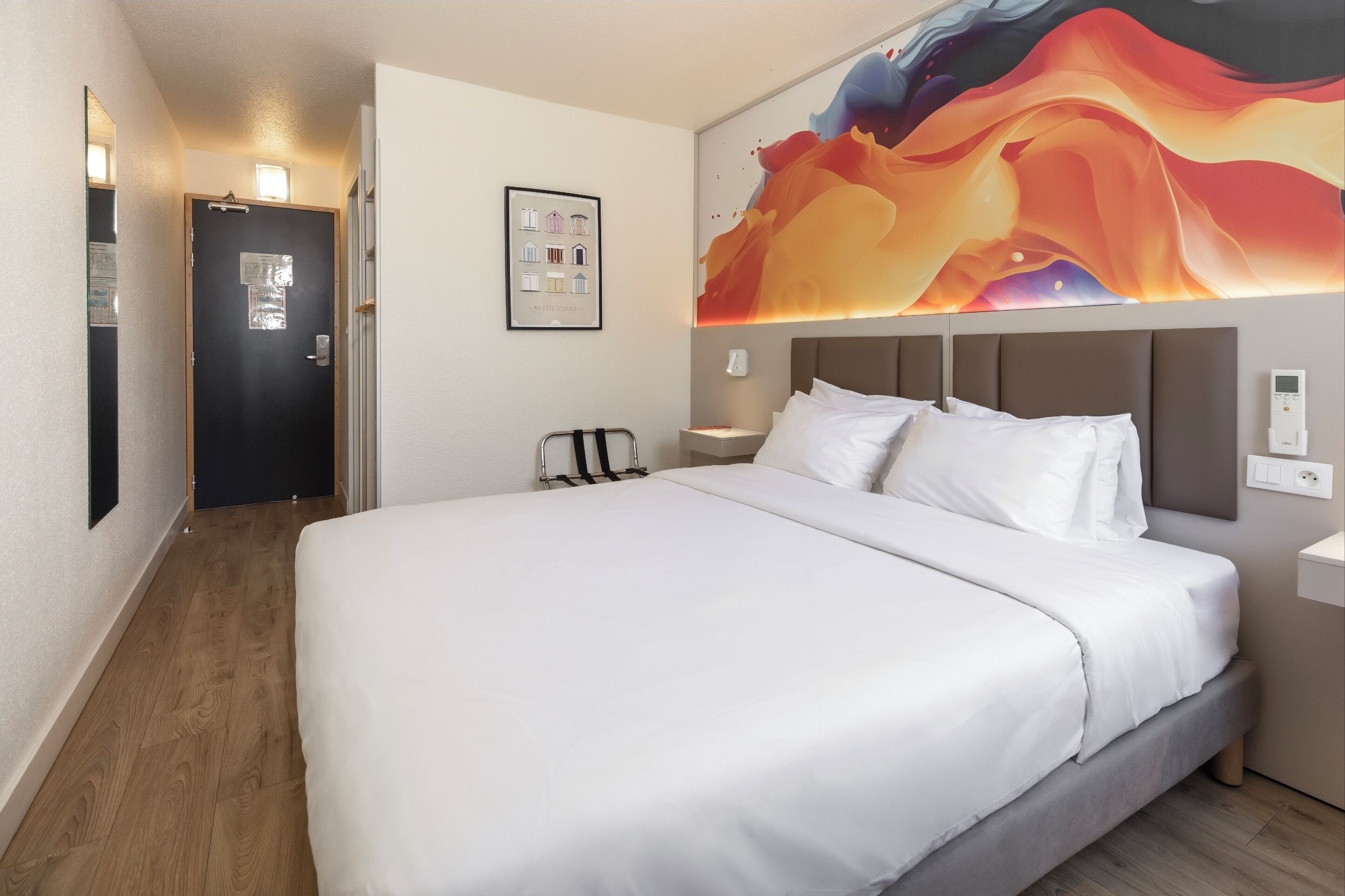 A hotel room with a large bed, a headboard, a wall-mounted artwork, a bedside table, and a door.