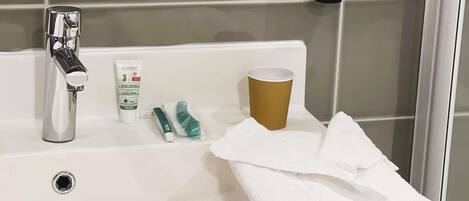 Un lavabo con grifo cromado, una botella de lavado Logodent para cabello, cuerpo y manos, un vaso de papel y una toalla blanca doblada.