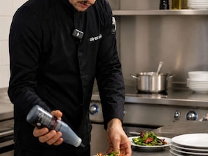 A chef in a black uniform with a name tag, preparing food in a kitchen with stainless steel appliances and a pot on the stove.