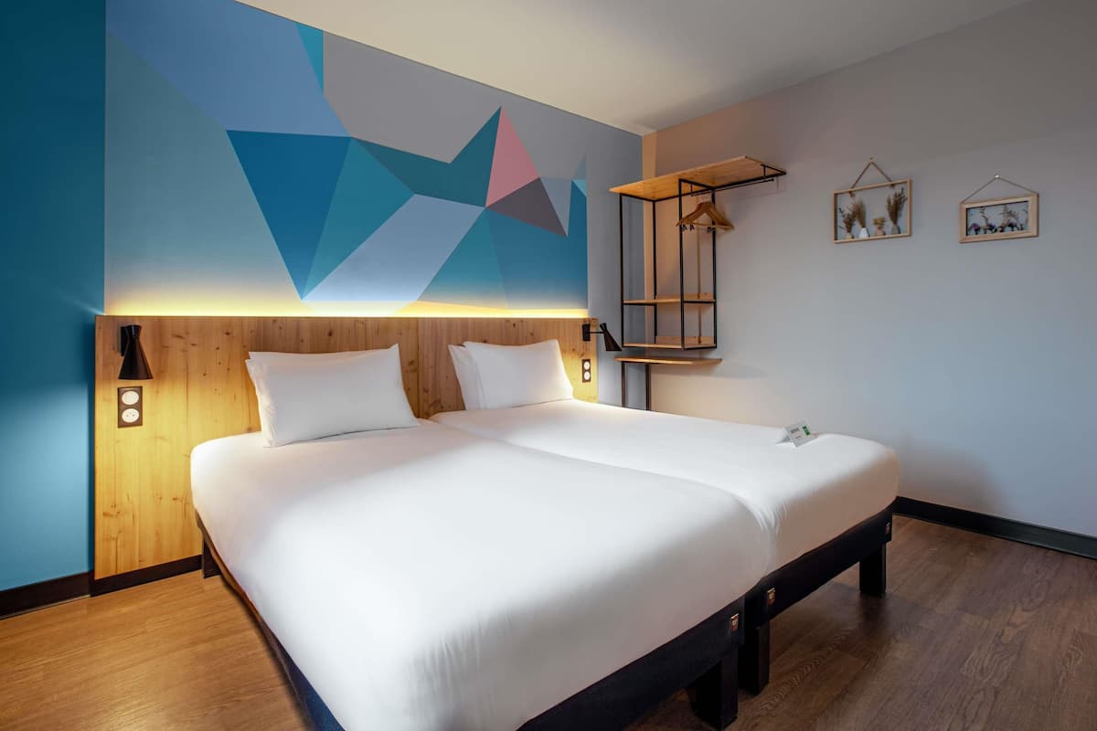 A modern hotel room with a large bed, wooden headboard, geometric wall art, bedside lamps, and a small shelf with decorative items.