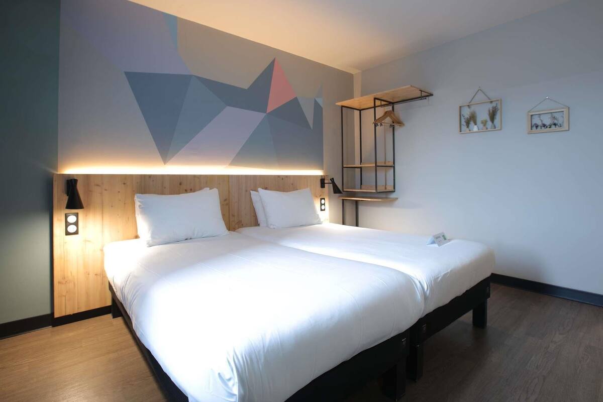 A modern hotel room with a large bed, a geometric wall mural, a bedside table with a lamp, and a small shelf with decorative items.