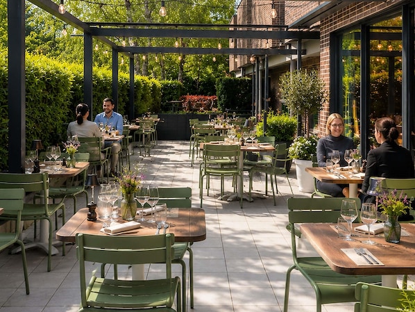 An outdoor dining area with green chairs and tables, surrounded by greenery and brick walls.
