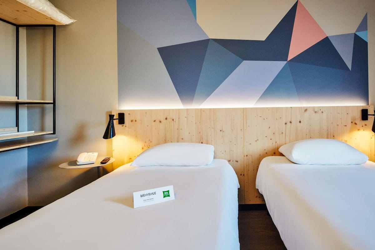 A hotel room with two beds, a wooden headboard, a geometric wall art, a bedside table with a lamp, and a welcome sign.