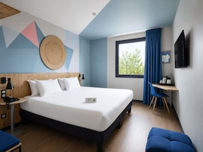 A modern hotel room with a bed, bedside table, lamp, and a desk with a chair.