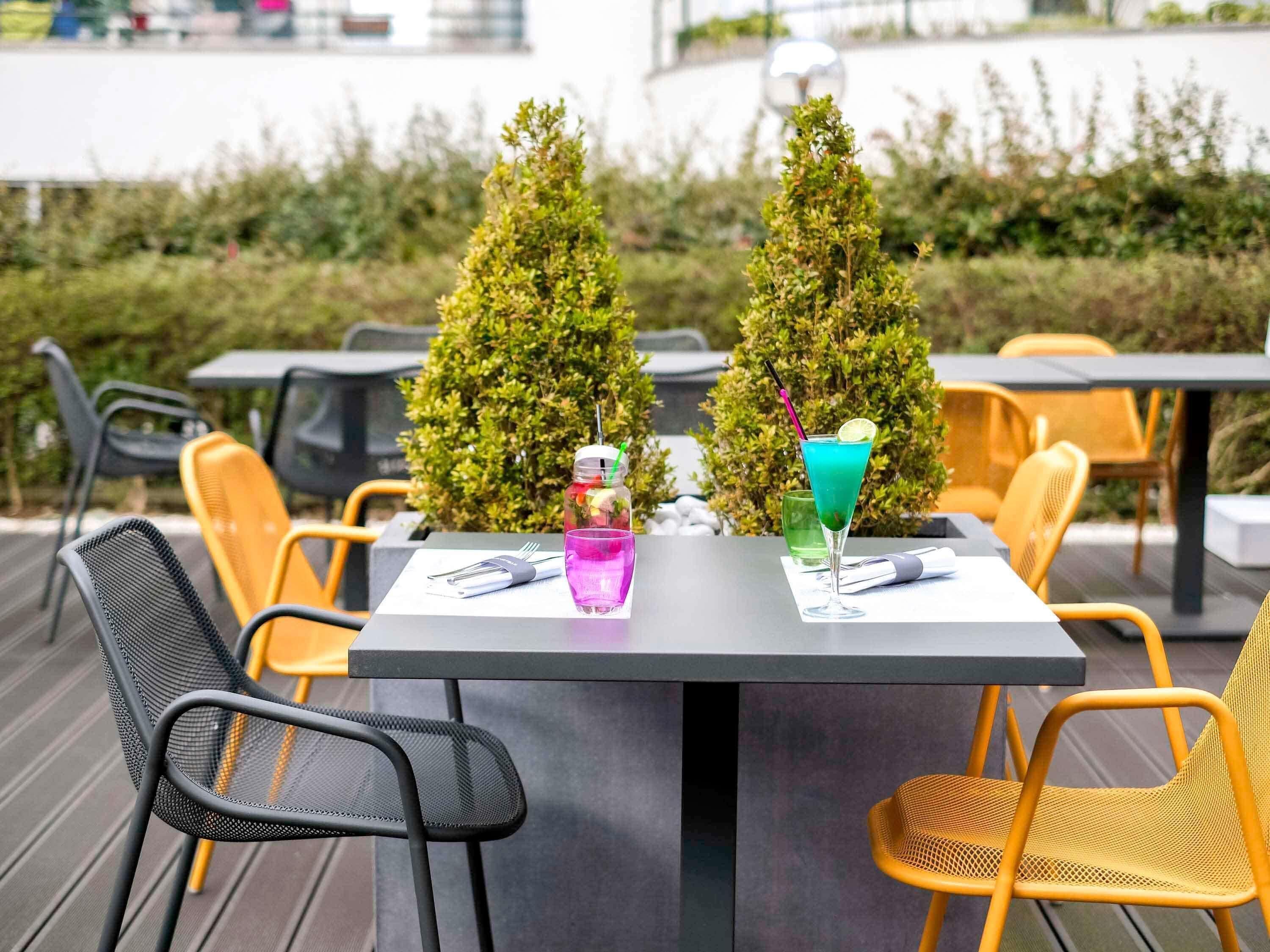 Un área de comedor al aire libre con mesas y sillas, dos árboles decorativos y una bebida sobre la mesa.