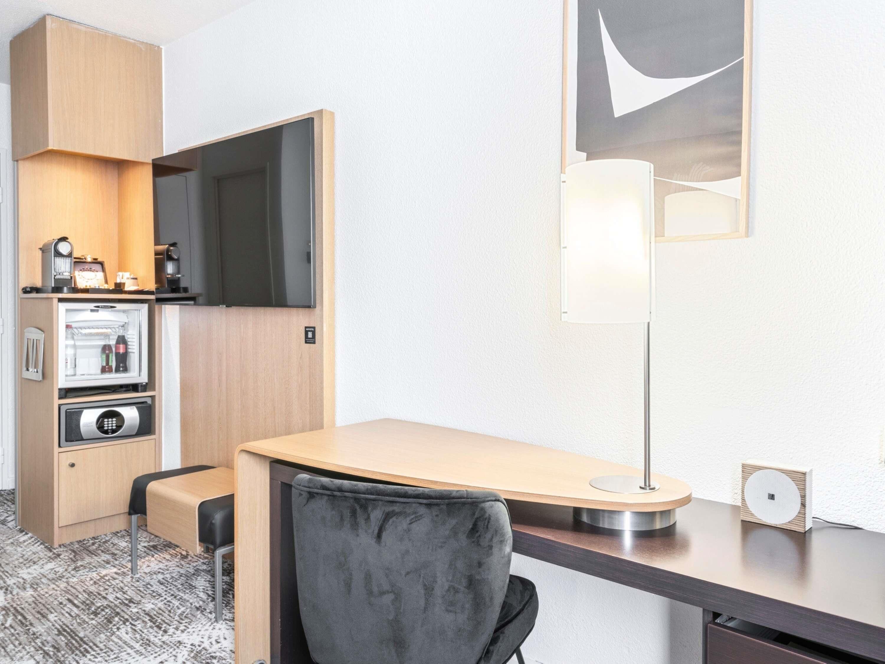 Een compacte hotelkamer met een kitchenette, een flatscreen televisie, een bureau met stoel en een moderne lamp.