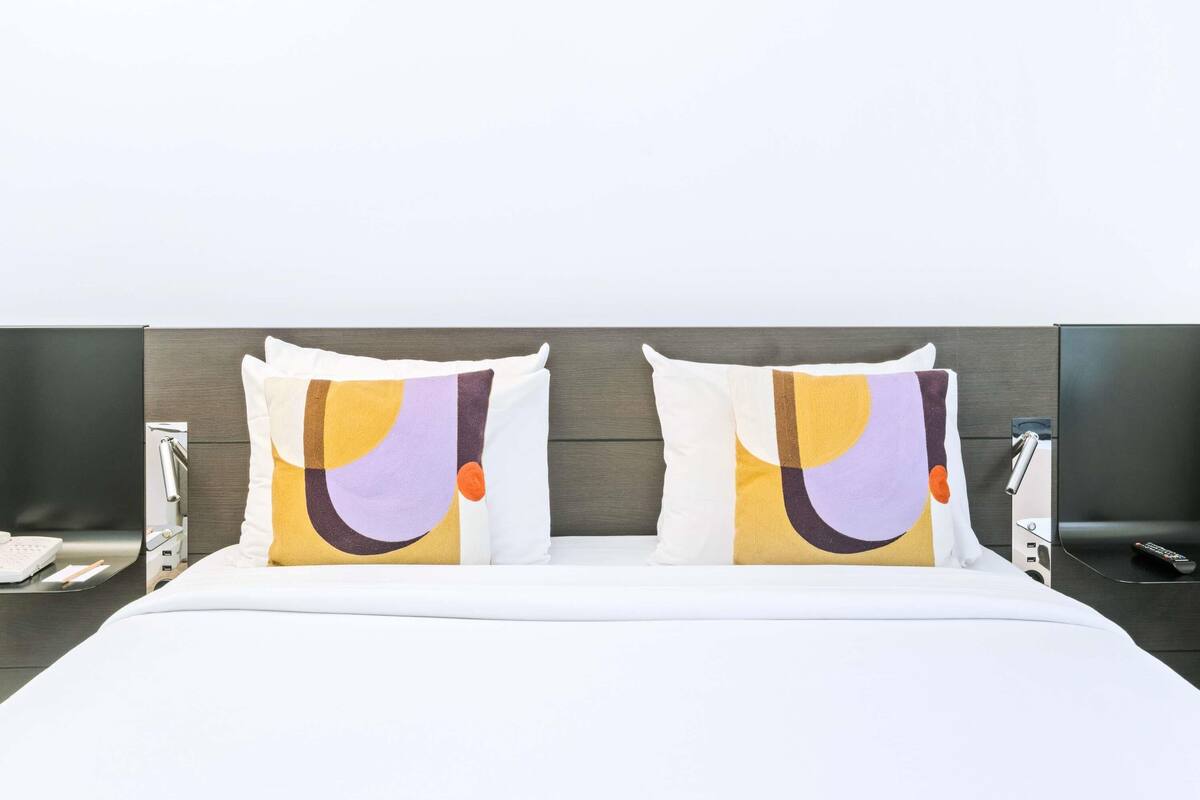 Habitación de hotel con una cama, dos almohadas de diseños abstractos, una mesita de noche con un teléfono y un televisor montado en la pared.