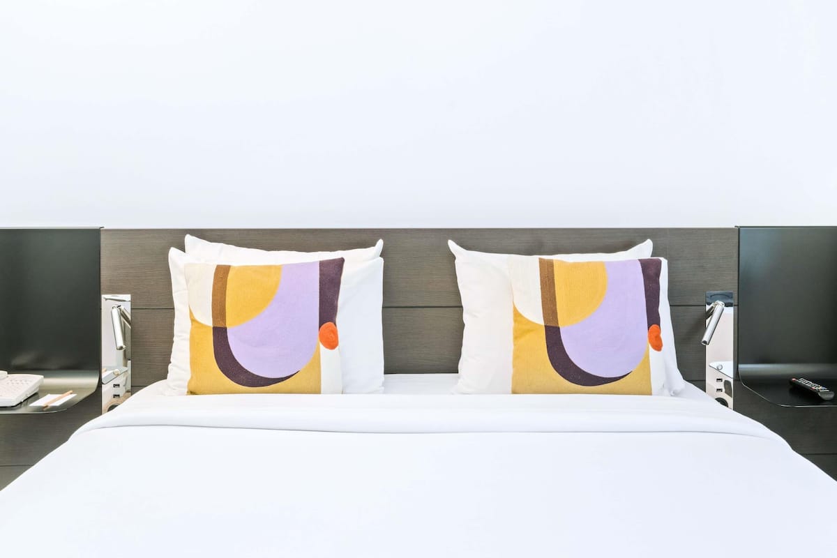 Une chambre d’hôtel avec un lit, deux oreillers aux motifs abstraits, une table de chevet avec un téléphone et un téléviseur fixé au mur.