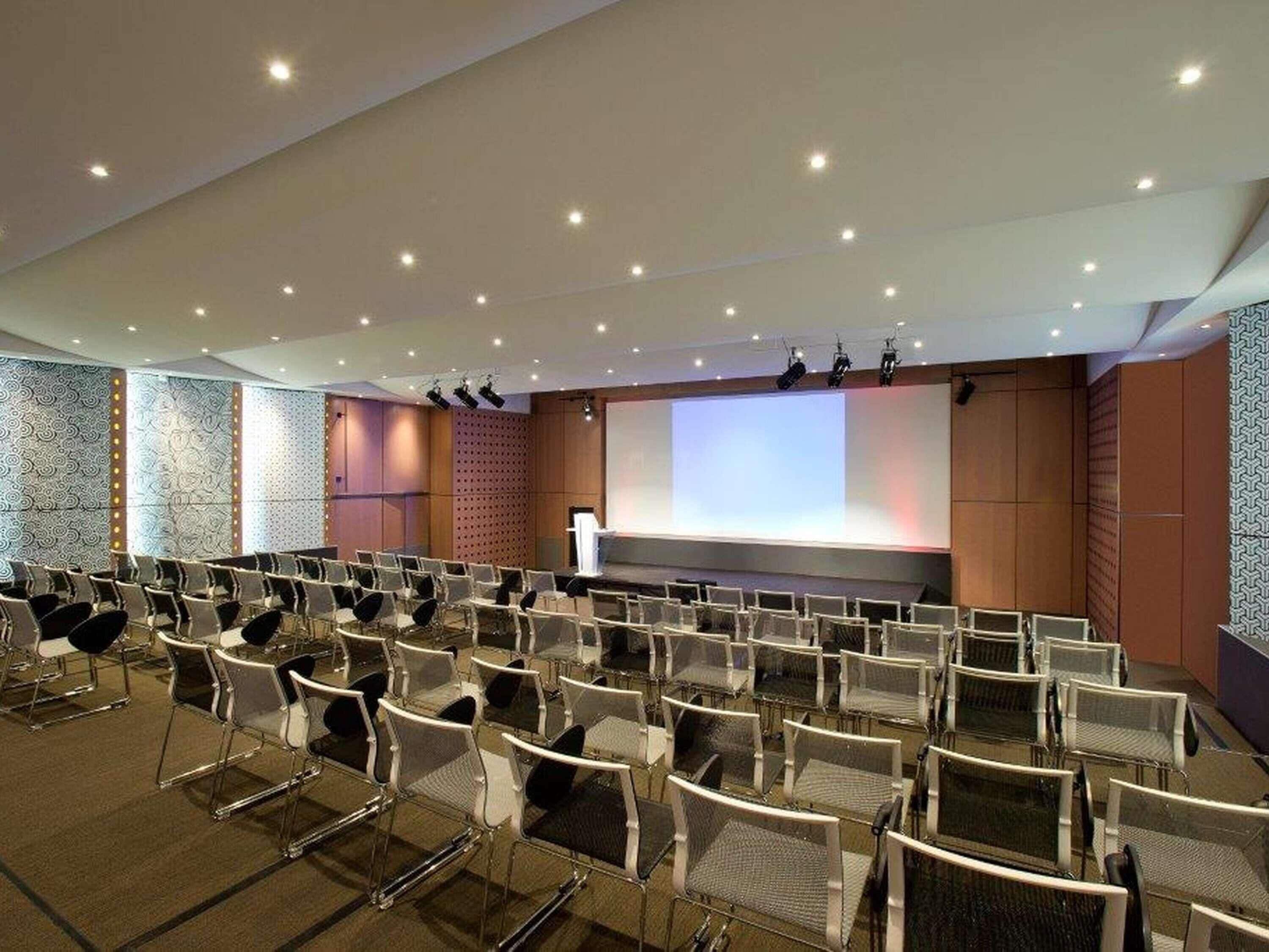 Una sala de conferències amb files de cadires, una pantalla gran i un escenari.