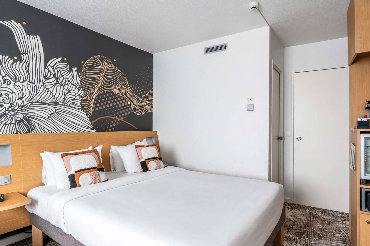 Una habitación de hotel moderna con una cama grande, una mesita de noche, un cabecero de madera, un mural en la pared, un armario y suelo alfombrado.