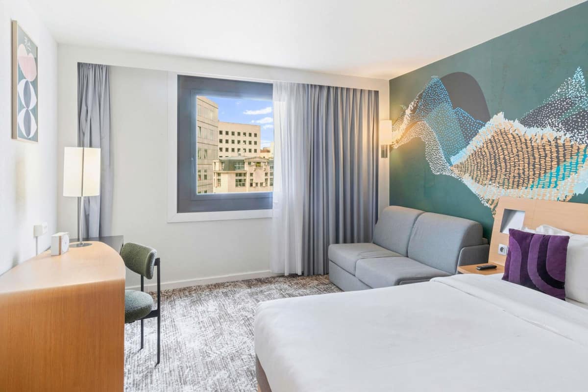 Habitación de hotel con cama, escritorio, silla, ventana con vista a edificios y una pared decorada con un relieve artístico.